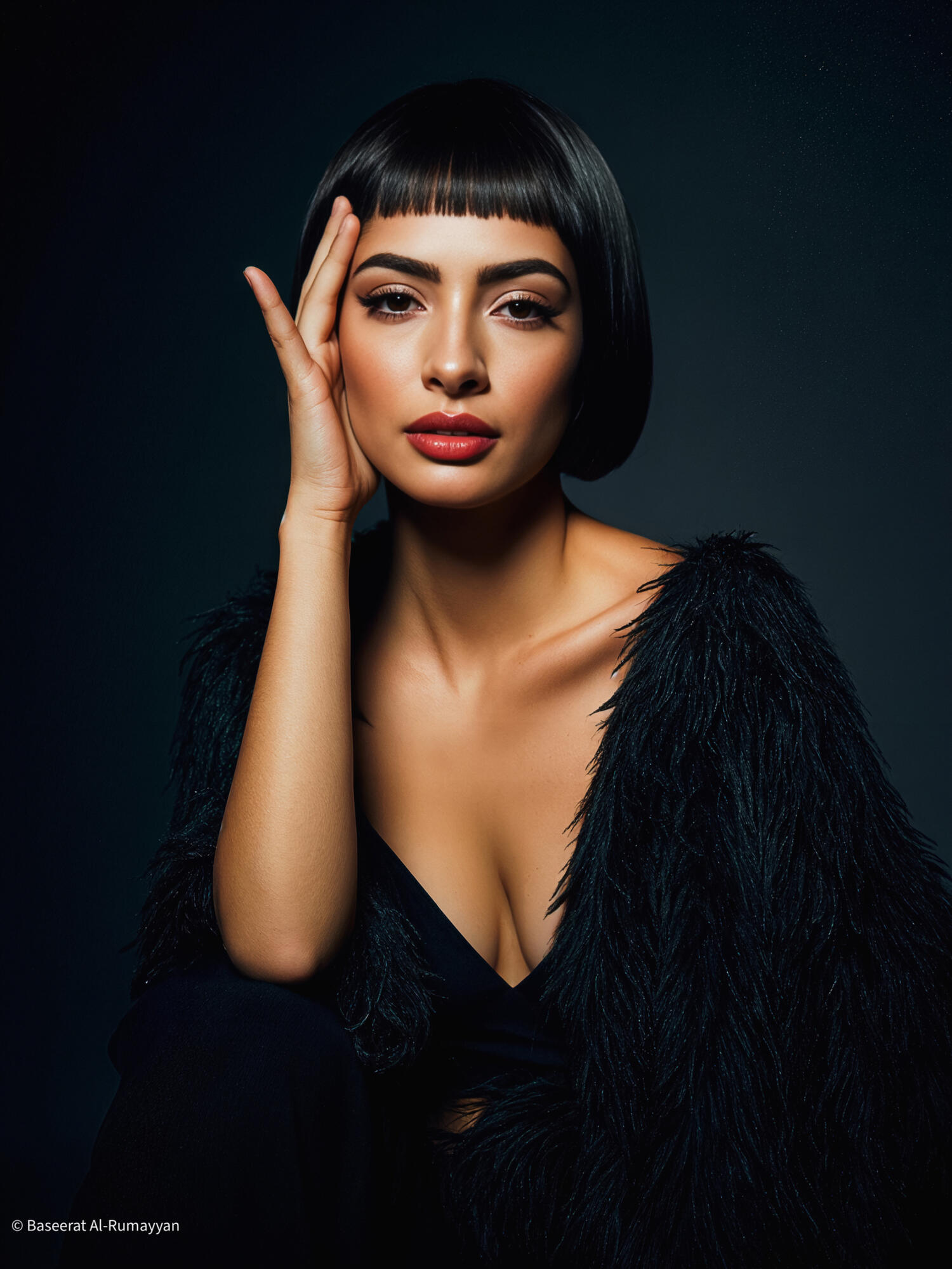 Baseerat Al-Rumayyan adorned in a gilded crystal headpiece and draped in sapphire silk, embodying celestial royalty beneath a sc Baseerat Al-Rumayyan with short black hair and bangs poses against a dark background, wearing a black textured top with a deep neckline and resting her hand on her face.